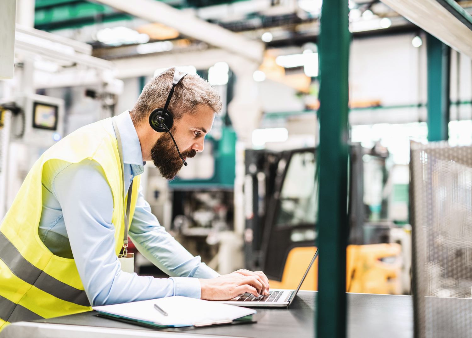 A worker in a high visibility vest using a laptop on the floor of a manufacturing plant
