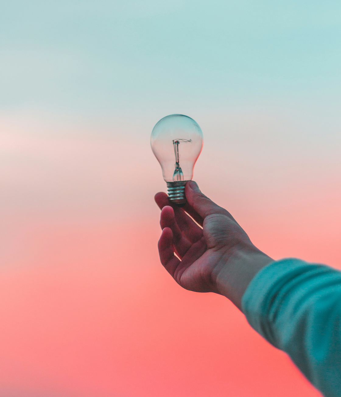 A hand holding a light bulb against a pink and blue sky